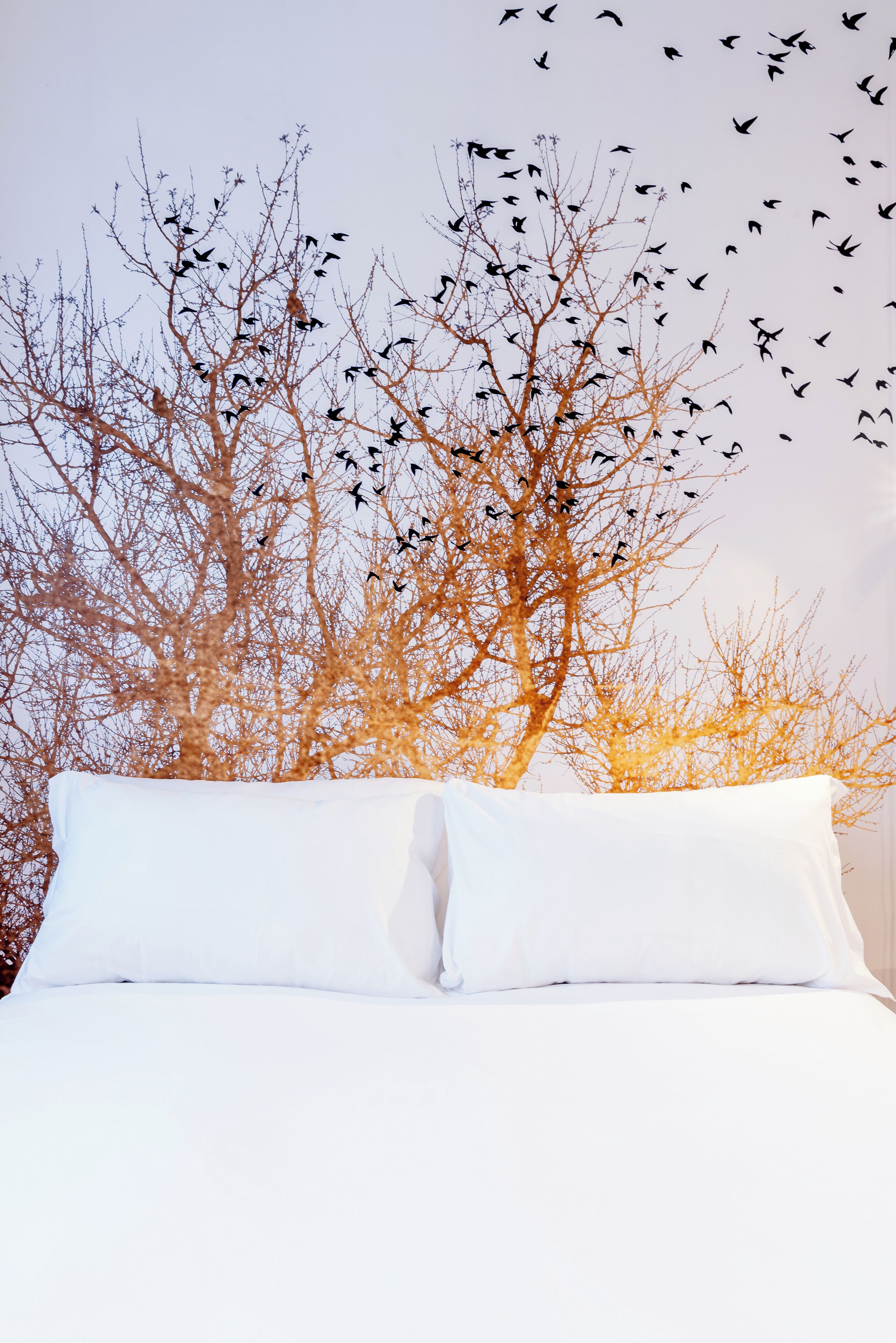 Un lit avec du linge de lit blanc et une vue sur un arbre aux branches nues, avec des oiseaux qui volent en arrière-plan.