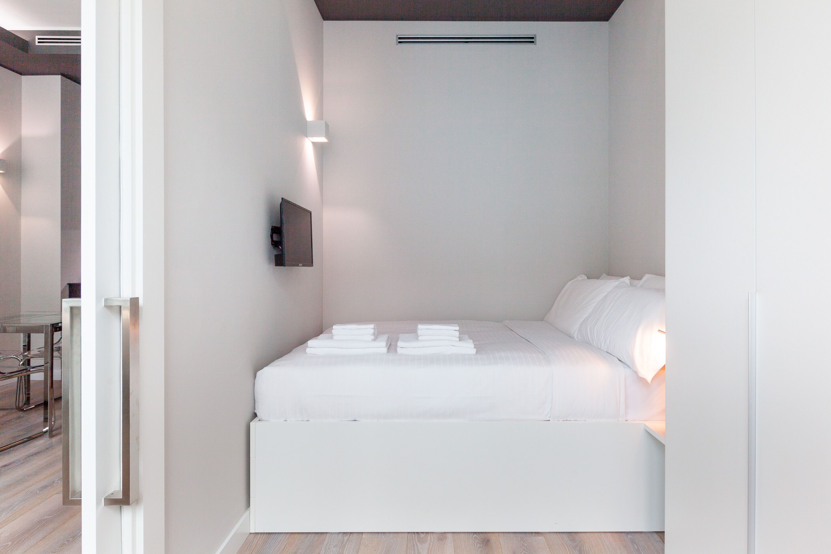 A modern, compact hotel room with a neatly made bed, a mounted flat-screen TV, and a small table with chairs.