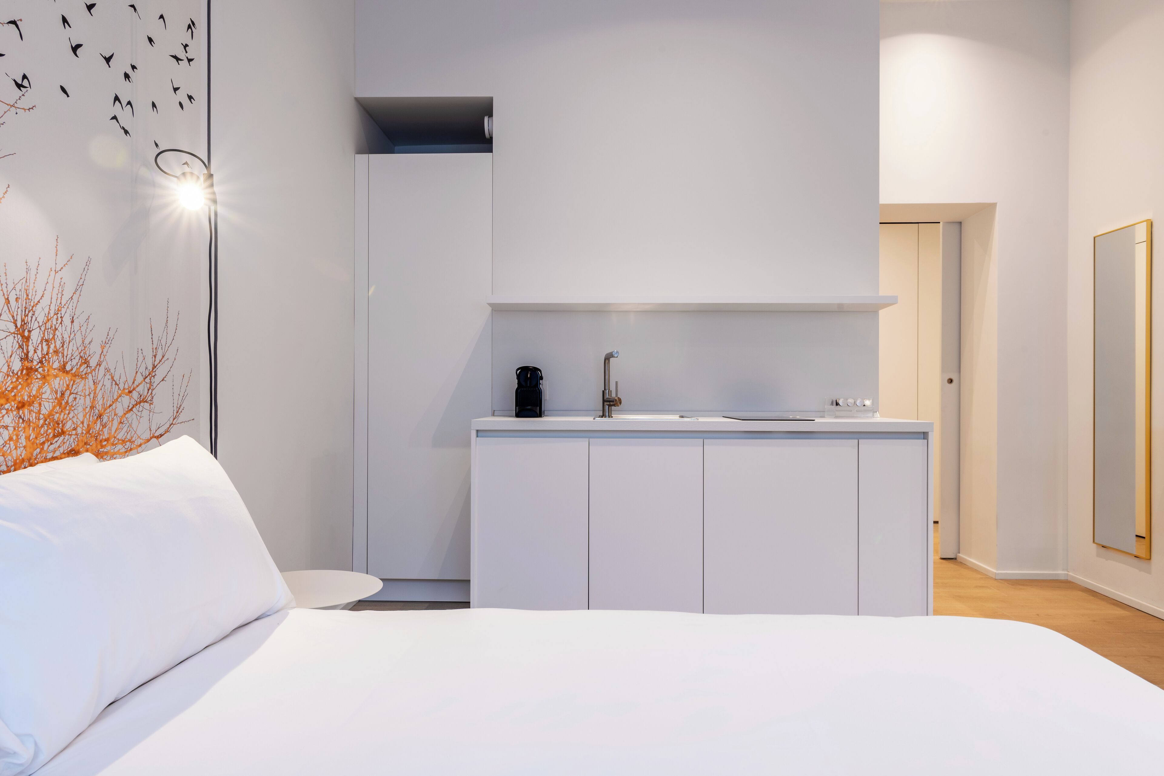 Un dormitorio moderno con cama, mesita de noche pequeña, armario empotrado y zona de kitchenette con lavabo y cafetera.