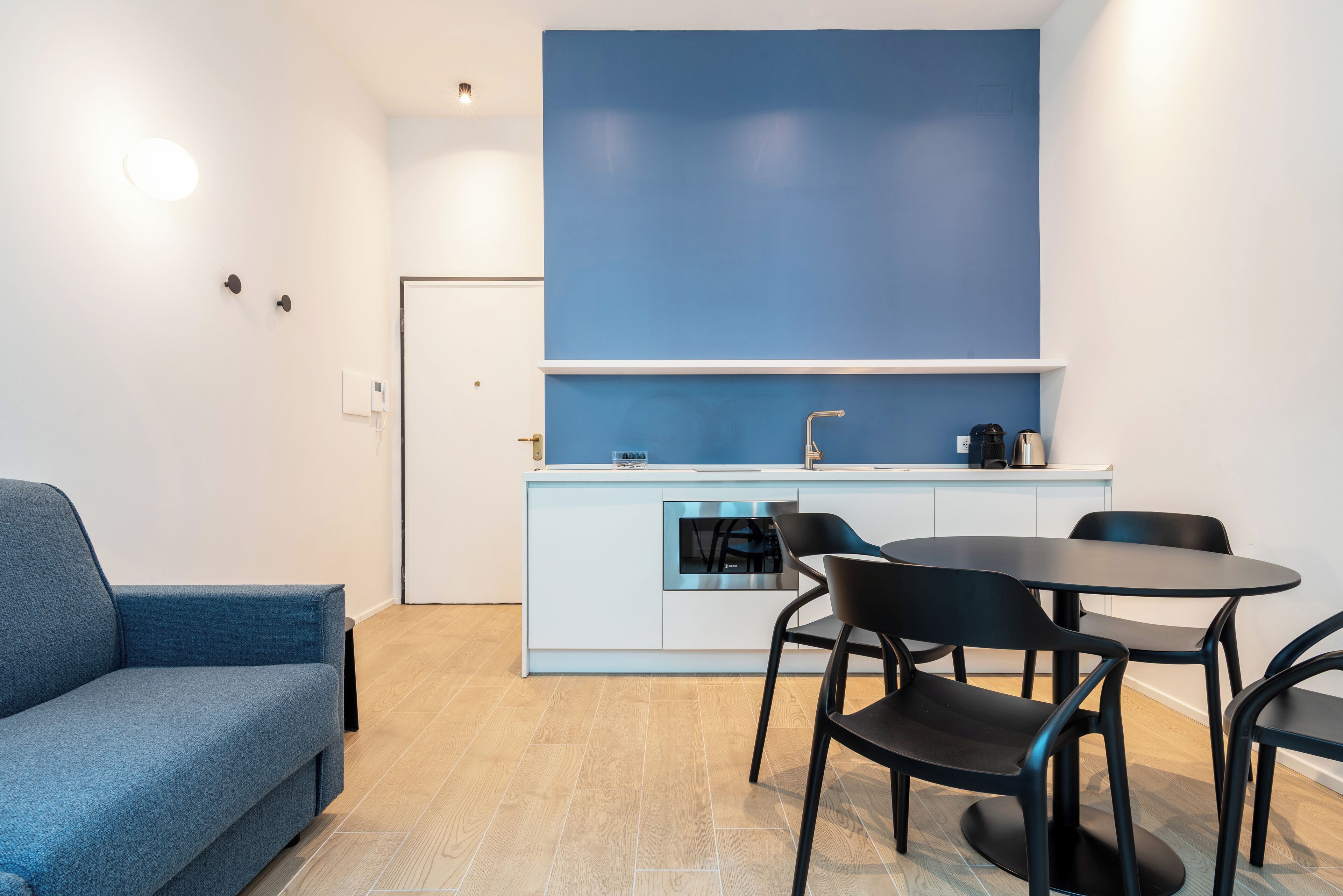 A compact kitchen with a blue accent wall, a dining area with black chairs, and a blue sofa.