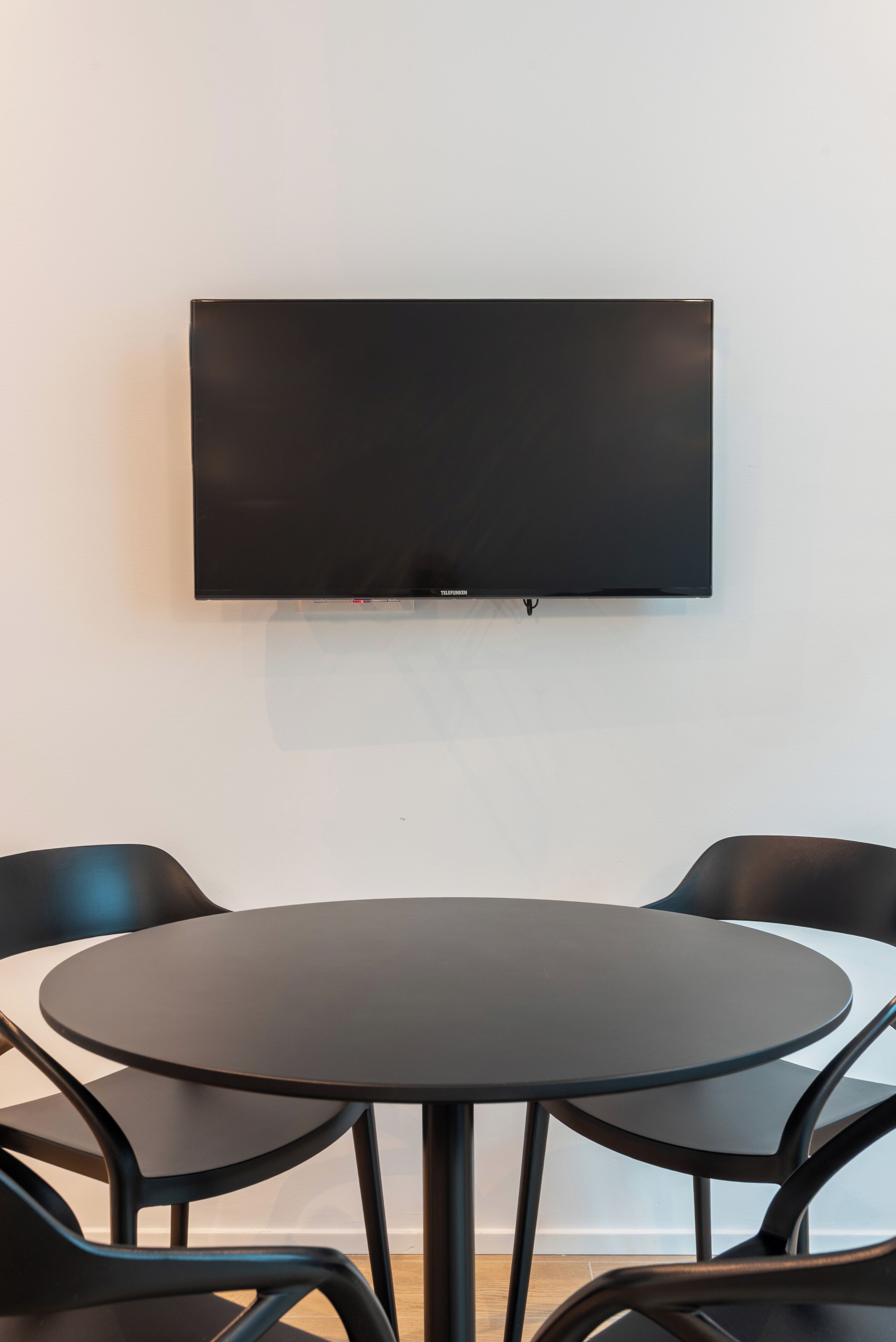 A modern room with a flat-screen TV mounted on the wall, a round table with four black chairs, and a light-colored wall.