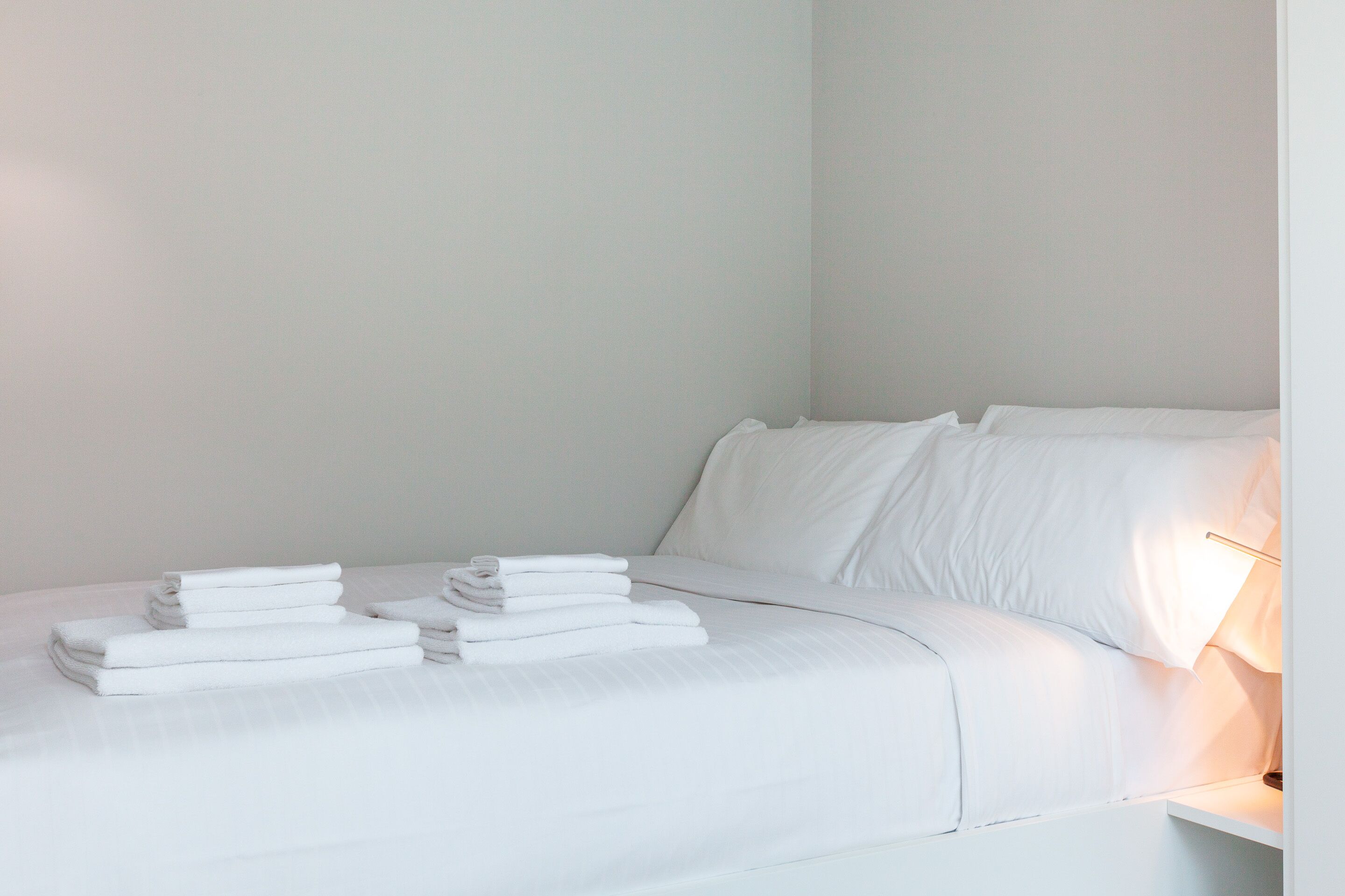 A neatly made bed with white linens and pillows, a bedside lamp, and a stack of folded towels.