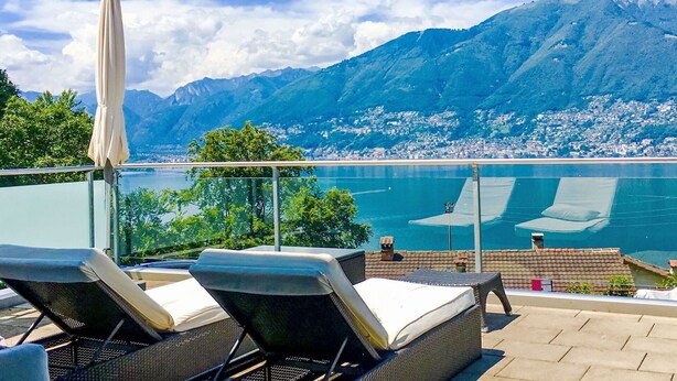 Une terrasse dotée de chaises longues et d’un parasol, offrant une vue sur un lac et des montagnes.