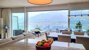 Une chambre d’hôtel moderne dotée d’une grande fenêtre offrant une vue sur la ville et les montagnes. On y trouve un coin repas avec une table et des chaises, ainsi qu’un panier de fruits posé sur la table.