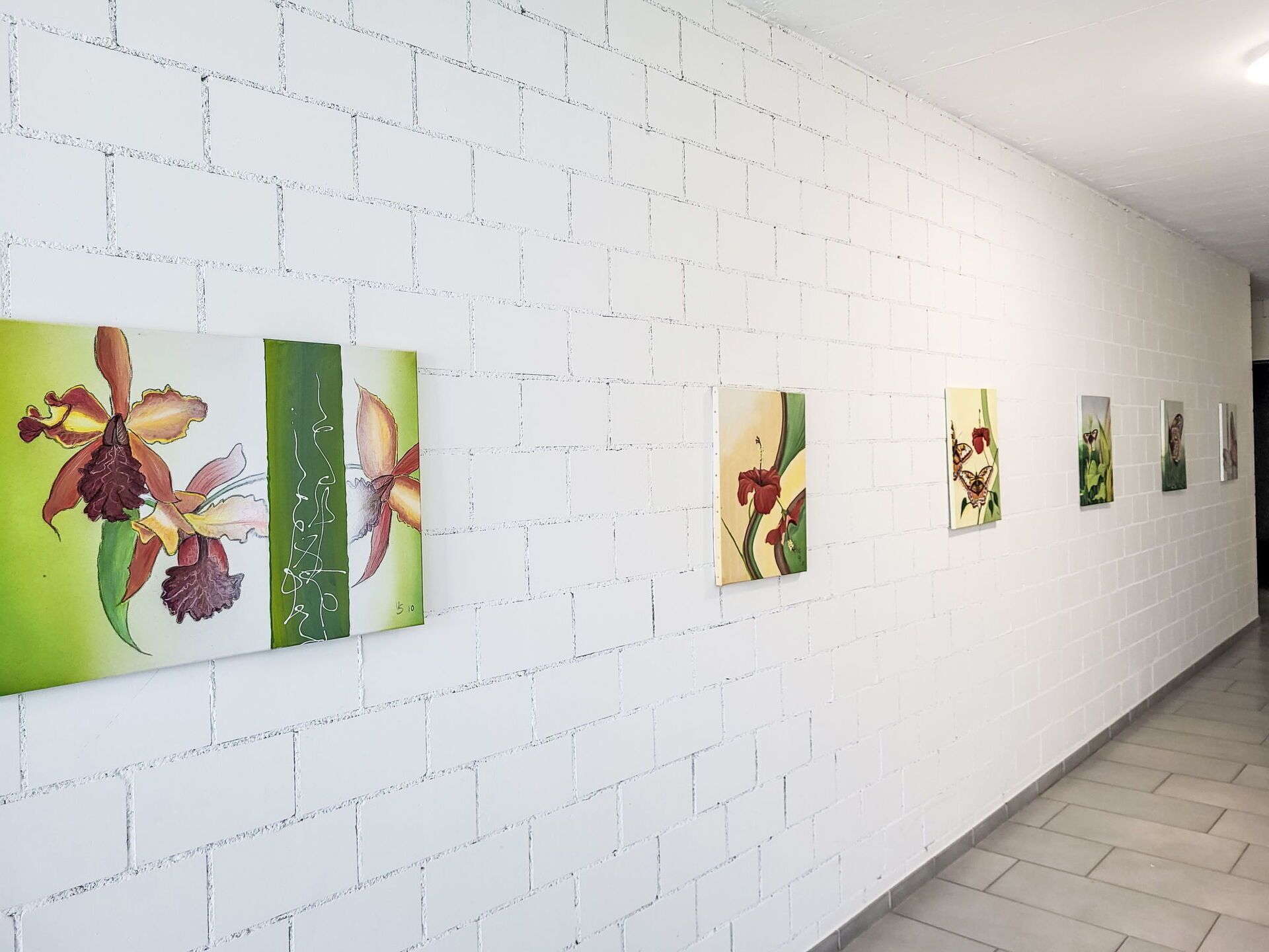 Un couloir aux murs en briques blanches et au sol carrelé, où sont exposées une série de peintures florales.