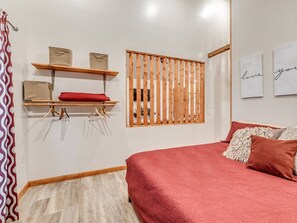 A bedroom with a bed, a wooden shelf with bags, a window with blinds, and a wall with framed text.