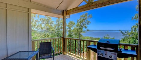 A covered outdoor area with a grill, a table, and a chair, overlooking a body of water.