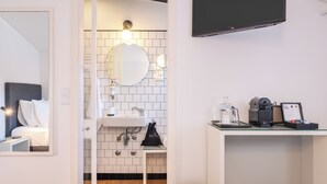Een moderne hotelbadkamer met een witte betegelde muur, een ronde spiegel, een wastafel, een koffieapparaat en een prullenbak.