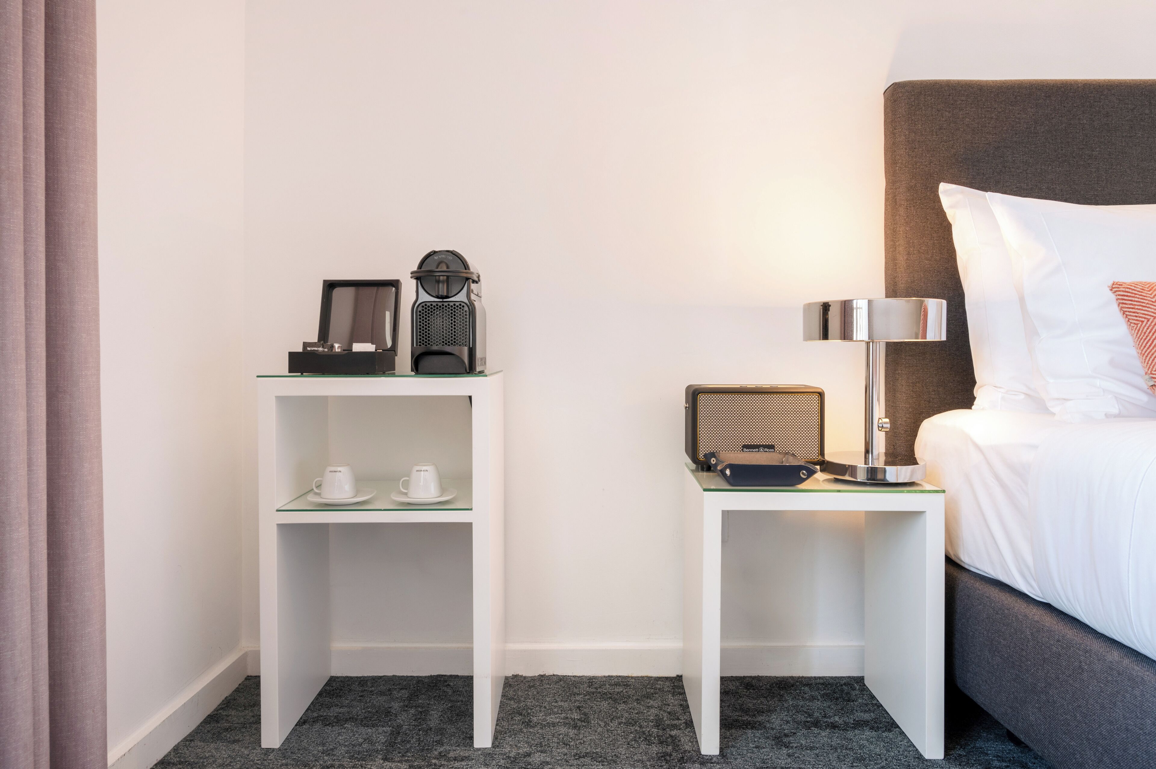 A hotel room with a bed, a nightstand with a lamp, a radio, and a shelf with cups.