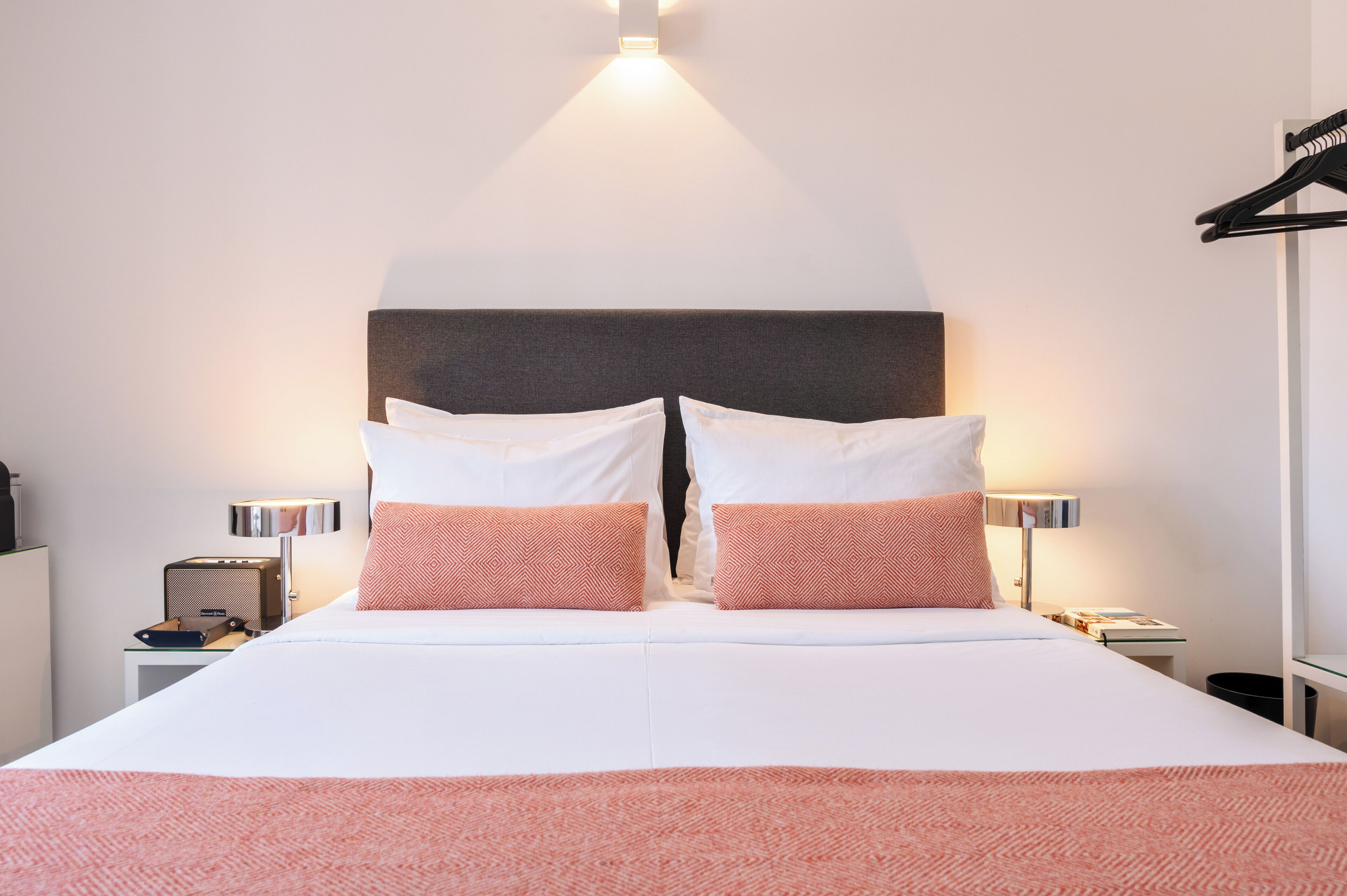 Una cama bien tendida con almohadillas blancas y rosadas, un cabecero oscuro y lámparas de noche.