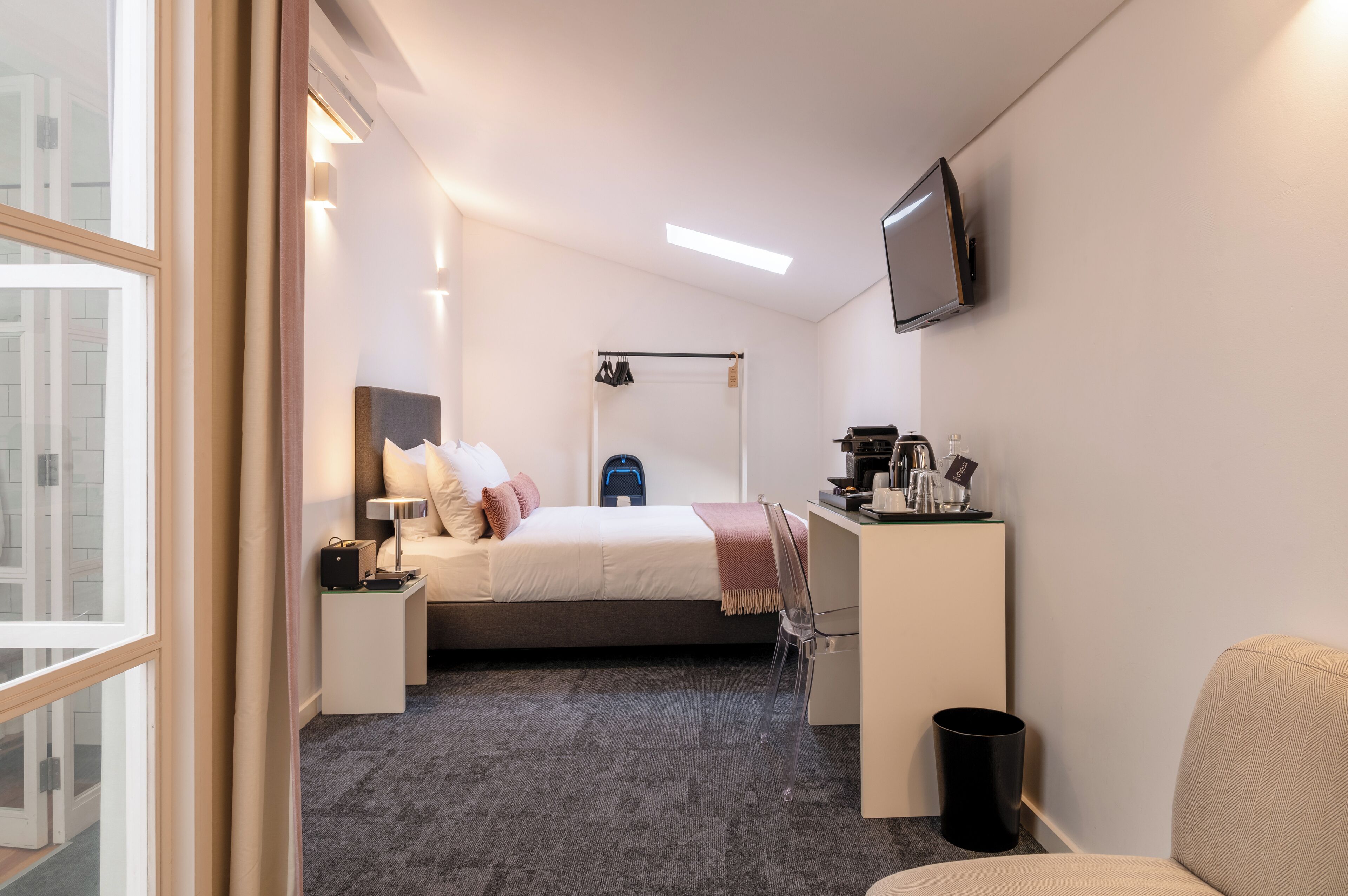 A modern hotel room with a bed, bedside table, chair, desk, TV, and a view of the city.