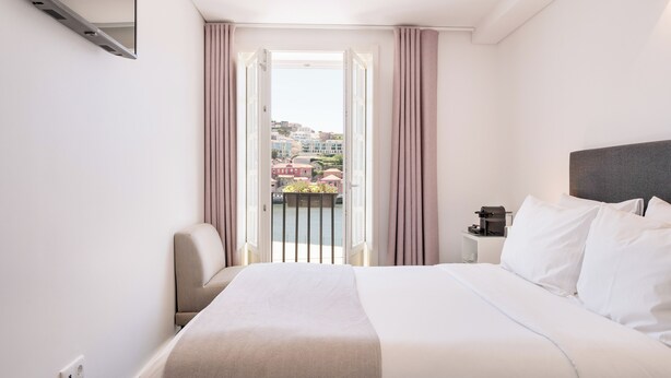 Hotelkamer met een bed, een stoel, een nachtkastje en uitzicht op gebouwen door een open deur.