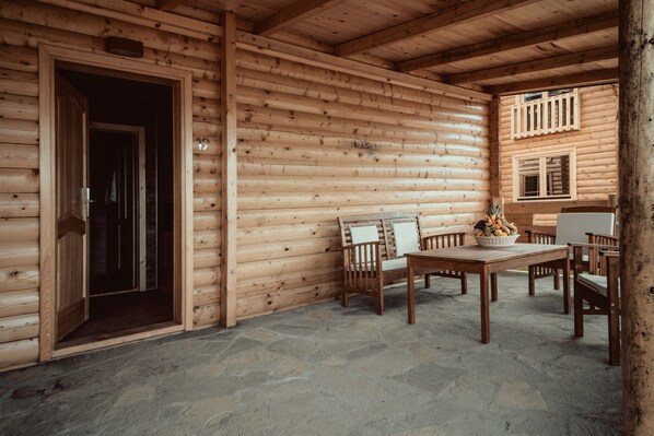 Interior de una cabaña de troncos con suelo de piedra, mobiliario de madera y una puerta que da a otra habitación.