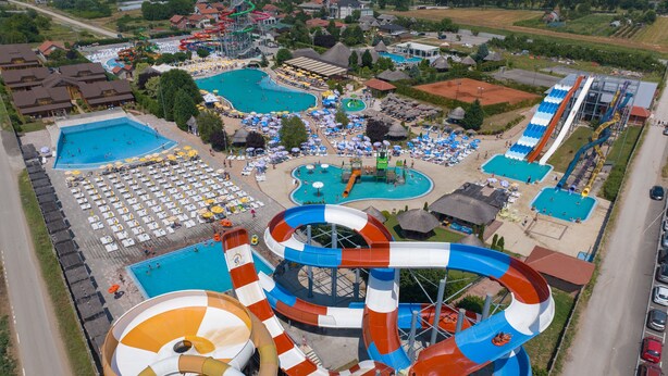 Vista aérea de un parque acuático con múltiples piscinas, un tobogán de agua grande y una zona de playa con sombrillas y sillas reclinables.