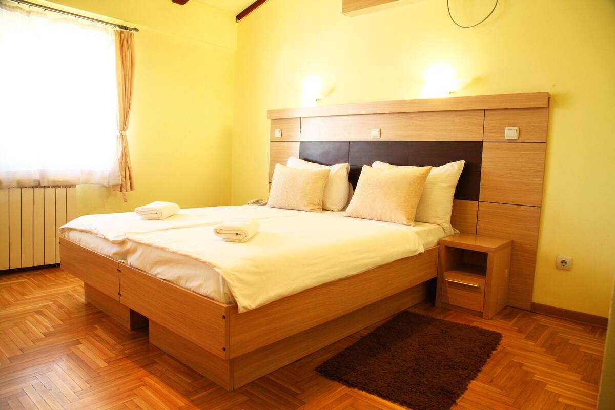 Cama bem arrumada com almofadas e toalha dobrada, mesa de cabeceira de madeira e tapete marrom sobre piso de madeira em quarto com paredes amarelas.