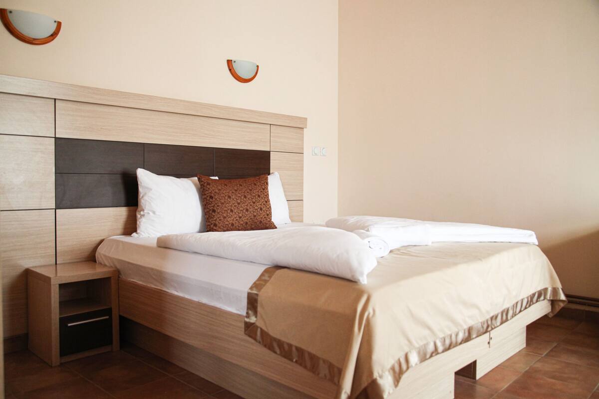 Uma cama bem arrumada com cabeceira de madeira, uma mesa de cabeceira e duas luminárias fixadas na parede.