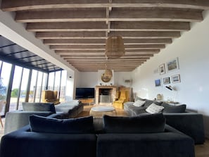 A modern living room with a high ceiling, built-in wooden beams, a fireplace, a television, and a large window offering a view of the outdoors.
