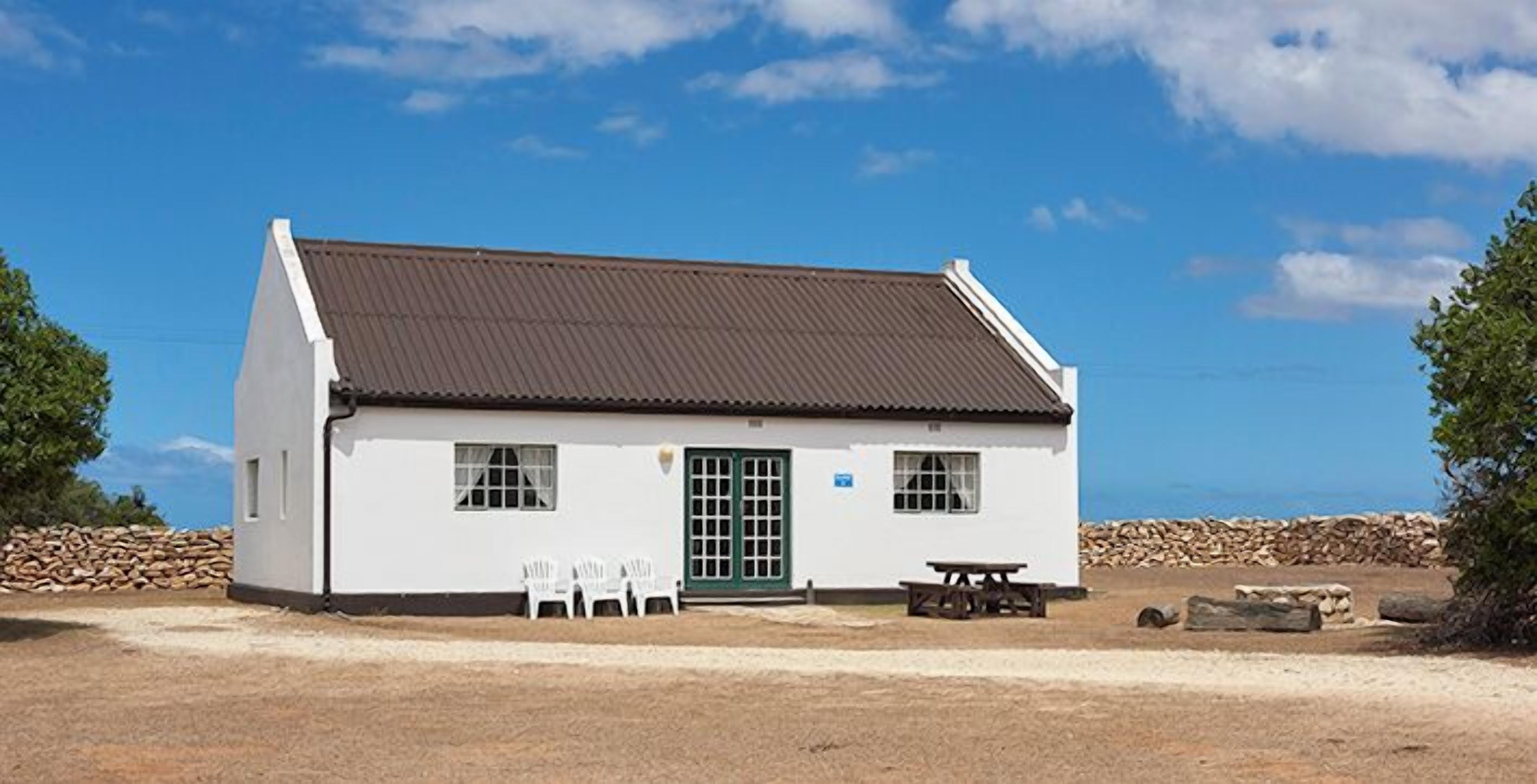 A white building with a brown roof, a door, and windows, with a gravel ground in front.