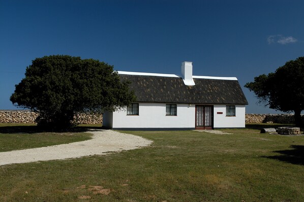 Una casa blanca con techo negro, una chimenea y un muro de piedra al fondo.