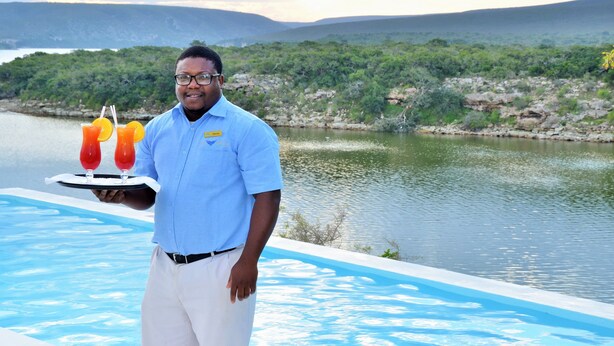 A person holding a tray with two drinks by a pool with a view of a lake and hills. - De Hoop Collection (Bredasdorp)