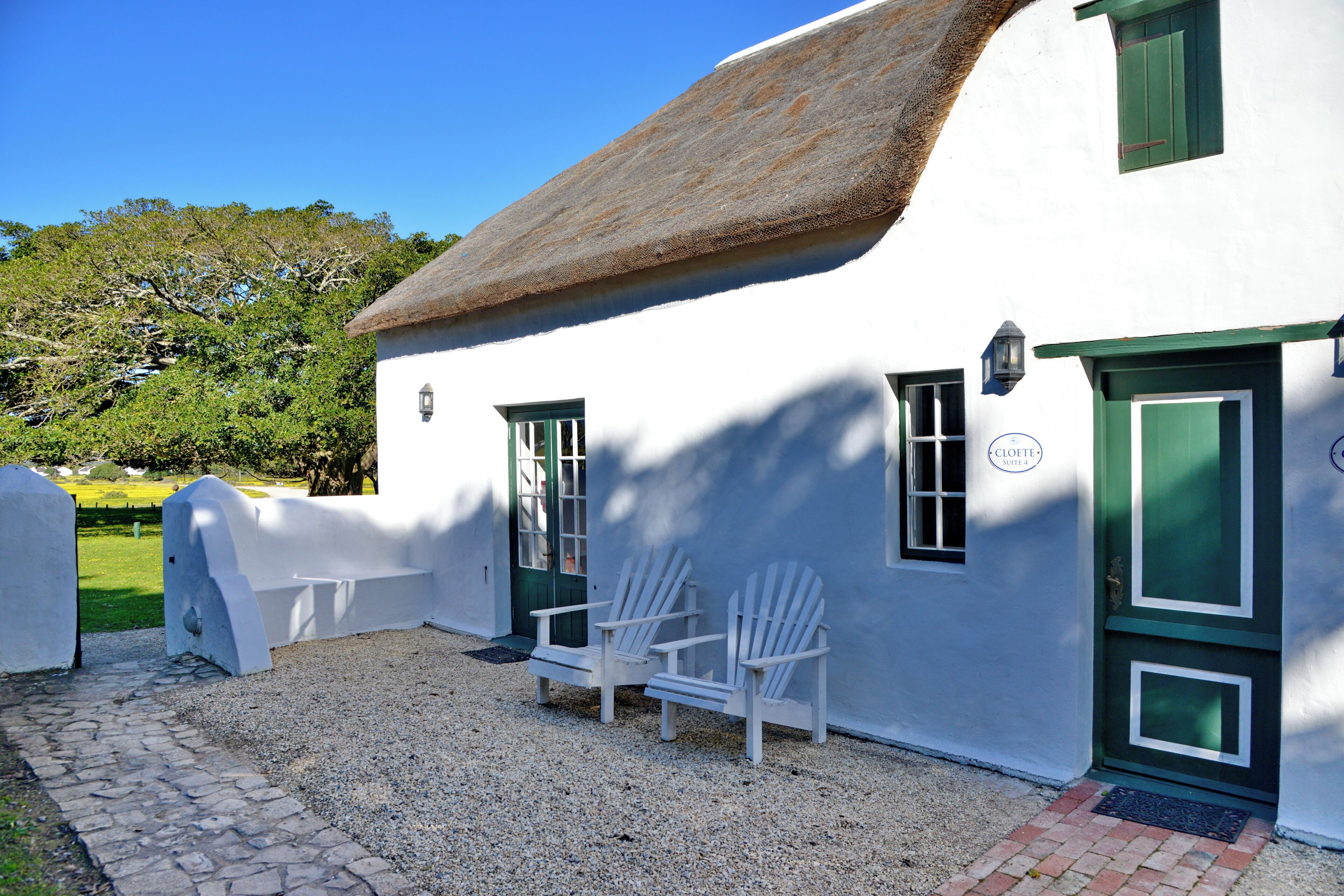 A white building with a thatched roof, two white chairs, and a gravel path.