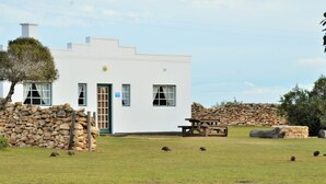 A white building with a stone wall, a wooden bench, and a tree. - De Hoop Collection (Bredasdorp)