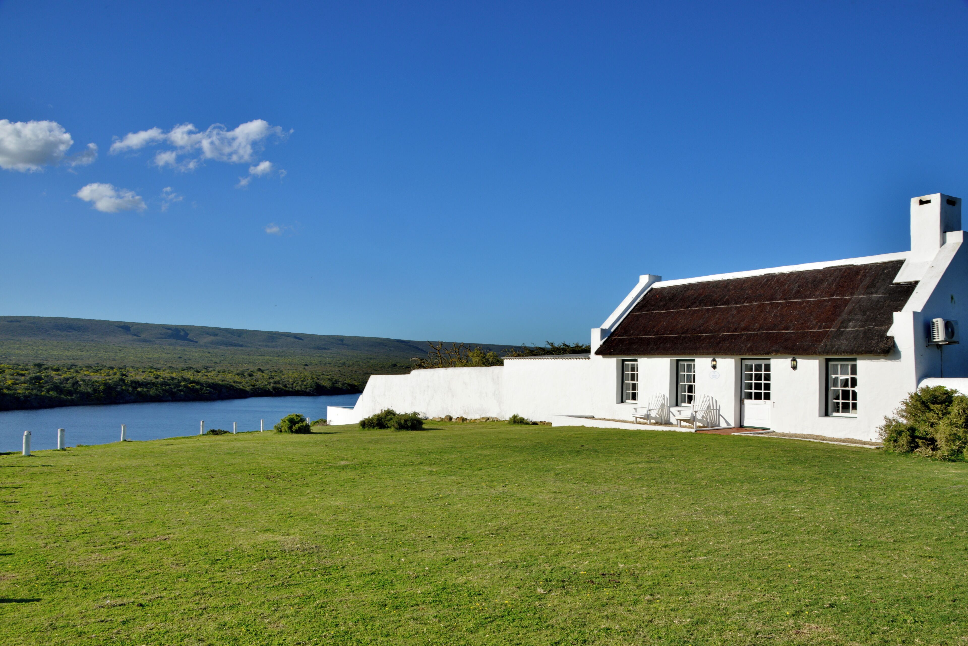 A white building with a dark roof, a grassy lawn, and a body of water in the background.