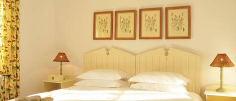 Habitación de hotel con cama, almohadas, mesita de noche, lámpara y pared decorada con ilustraciones botánicas enmarcadas.