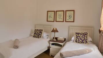 A hotel room with two single beds, a nightstand with a lamp, and three framed botanical illustrations on the wall.
