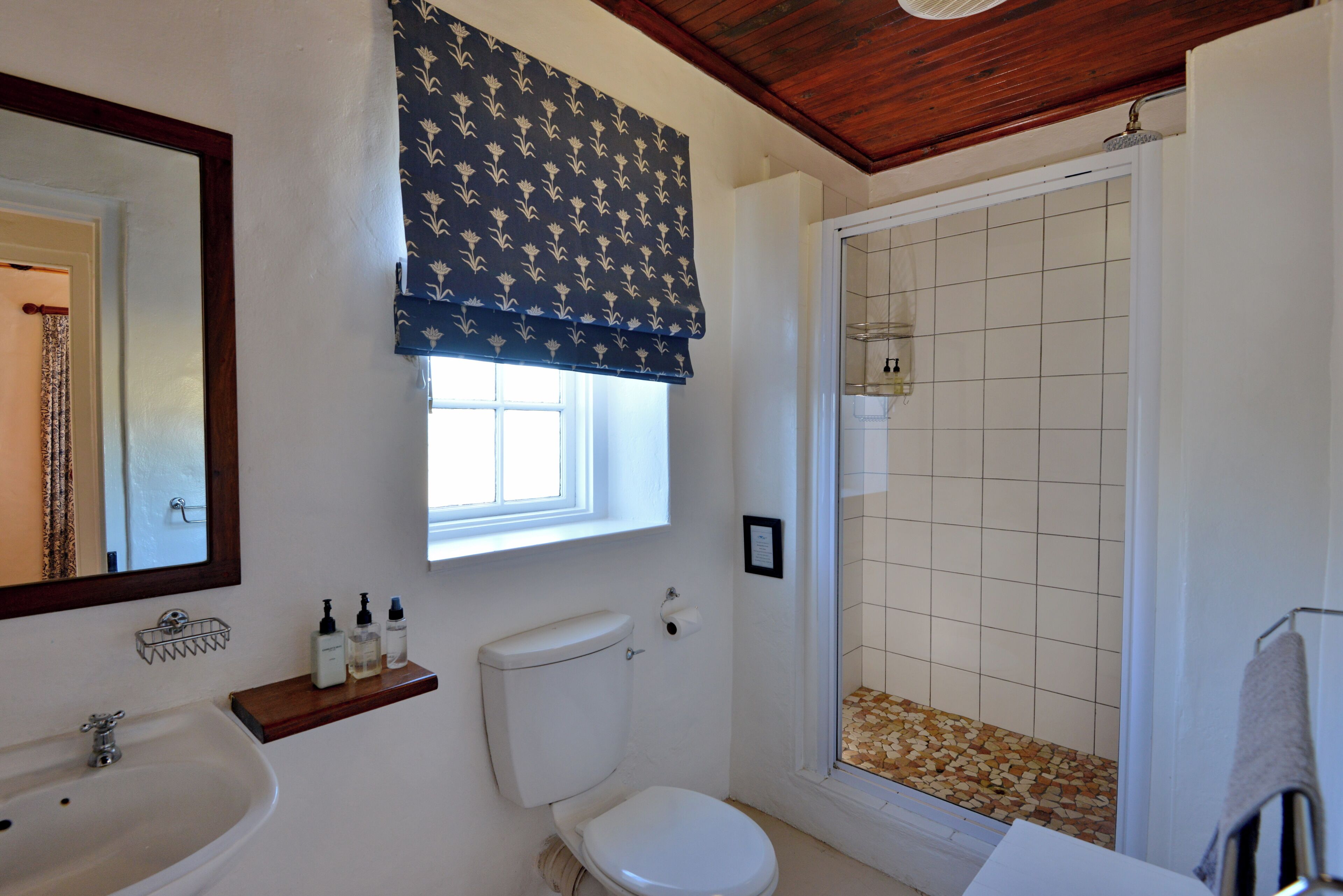 A bathroom with a white toilet, a sink with a wooden shelf, a mirror, and a shower with a tiled floor.