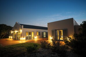 Una casa moderna de dos pisos con amplios ventanales, un patio cubierto y exteriores bien iluminados al anochecer.