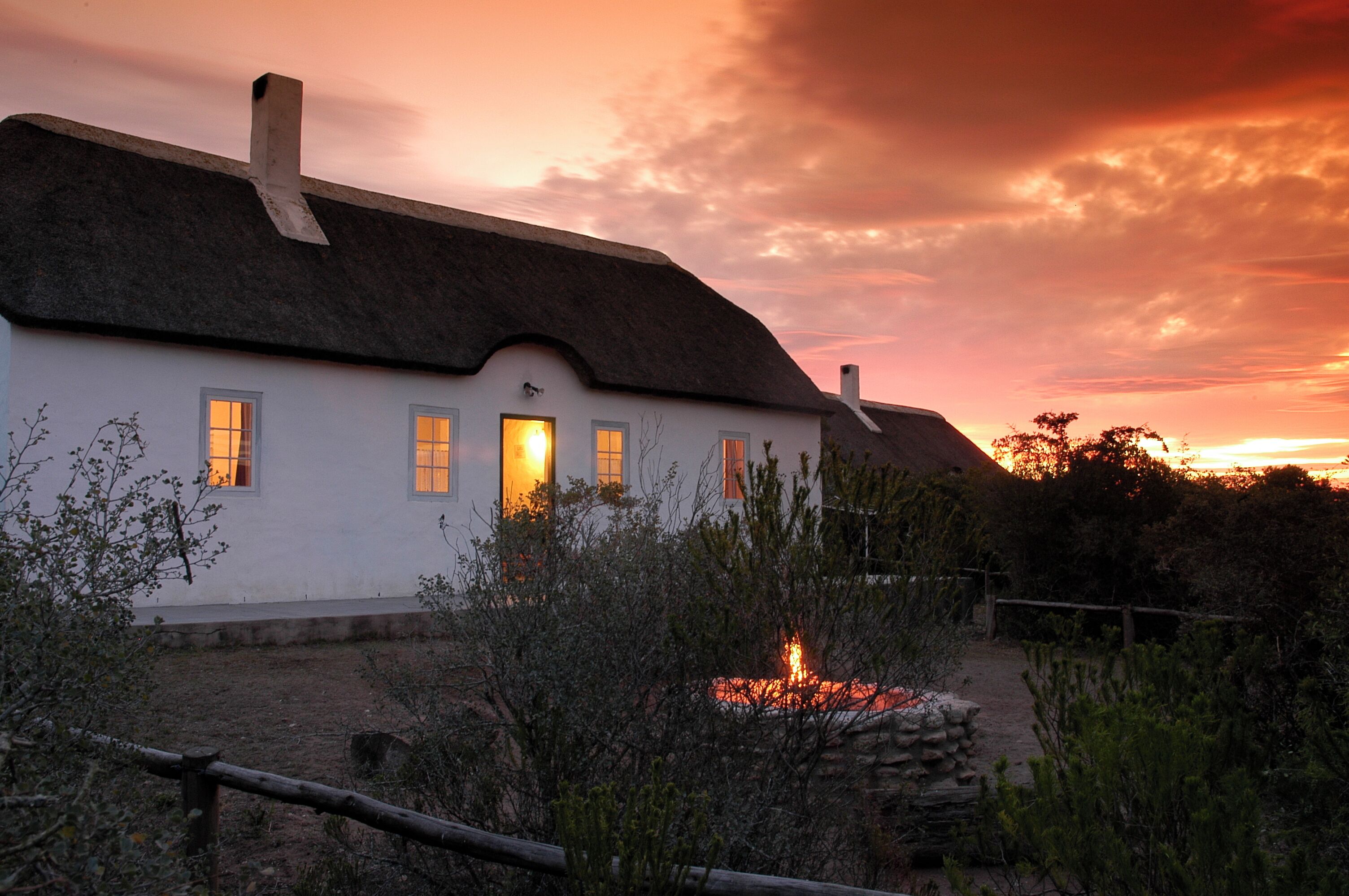A white building with a thatched roof, multiple windows, and a fire in the foreground.