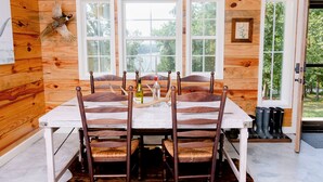 A wooden cabin with a dining table set for six, surrounded by wooden chairs. There are three windows with white frames, a wooden door, and a mounted bird on the wall.