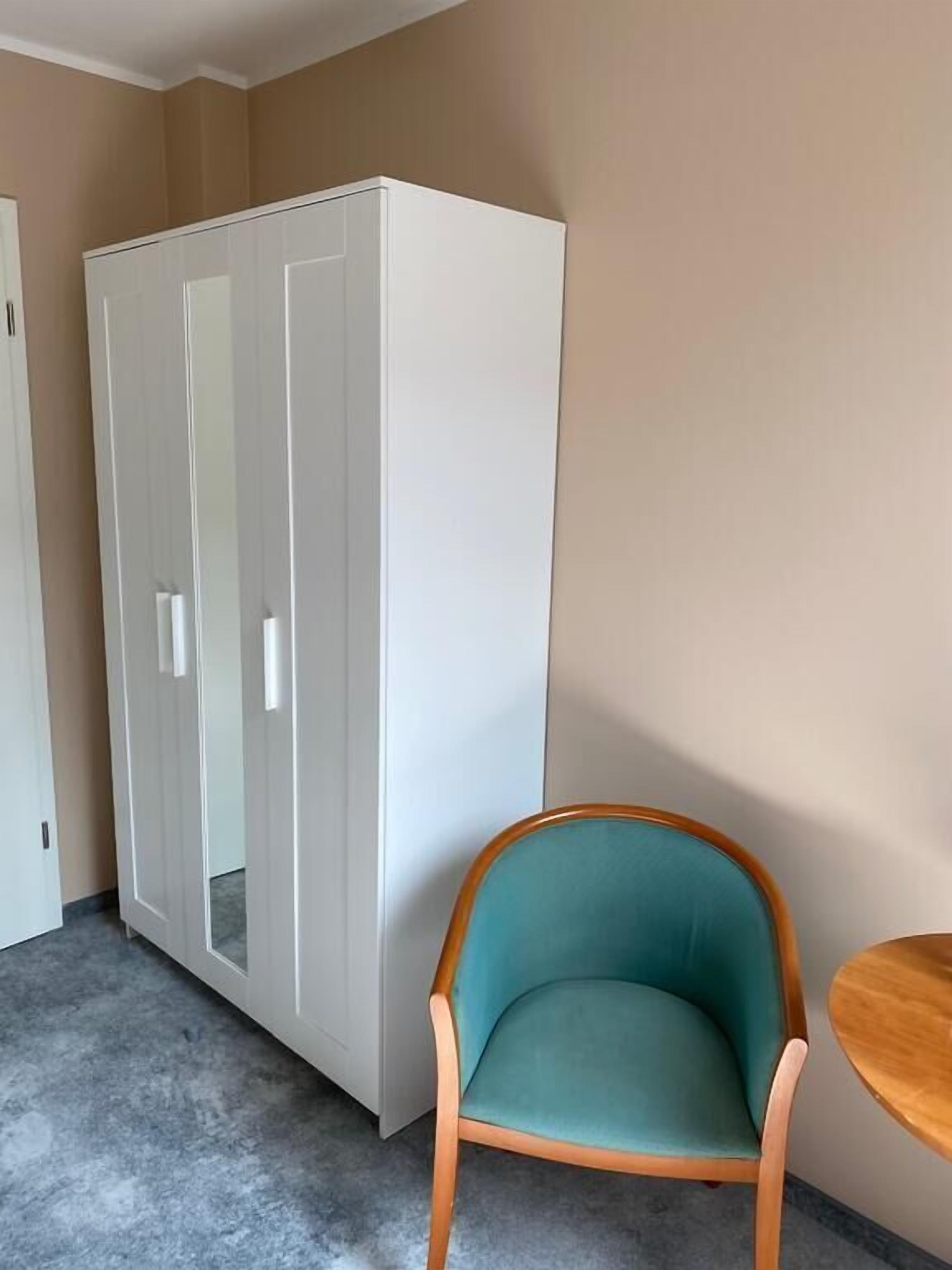 A room with a white wardrobe, a teal chair, and a wooden table.