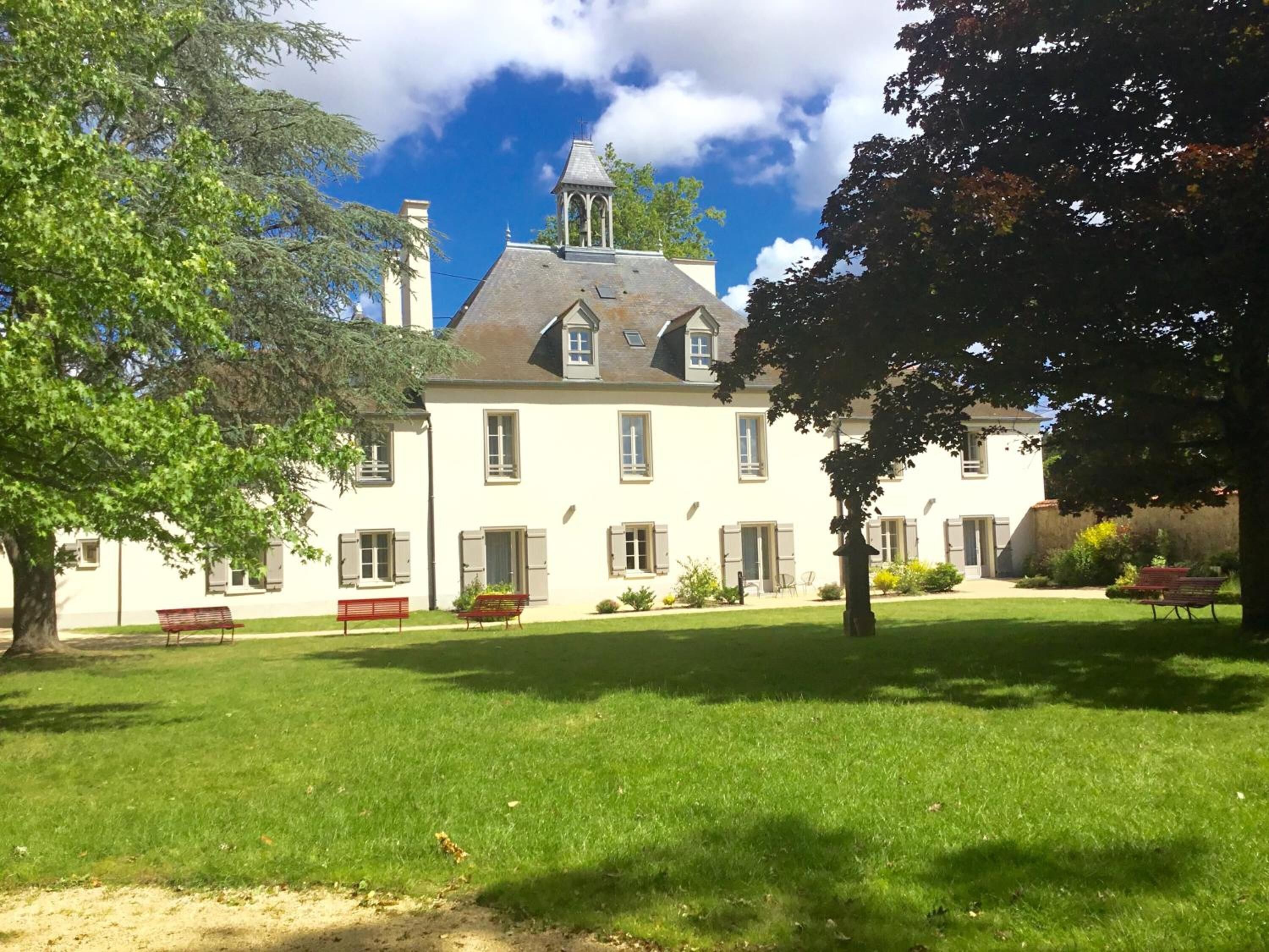 A large, white building with a steeply pitched roof and a small tower. There is a well-maintained lawn in front of the building with a few benches and a tree.