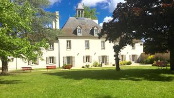 A large, white building with a steeply pitched roof and a small tower. There is a well-maintained lawn in front of the building with a few benches and a tree.