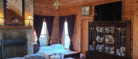 Une chambre confortable, aménagée dans le style d’un chalet, avec une cheminée en pierre, un lit placé près d’une grande fenêtre, une table à manger avec des chaises et un buffet.