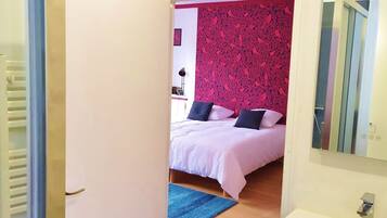A bedroom with a bed, a blue rug, a white wall, and a bathroom with a sink and mirror.