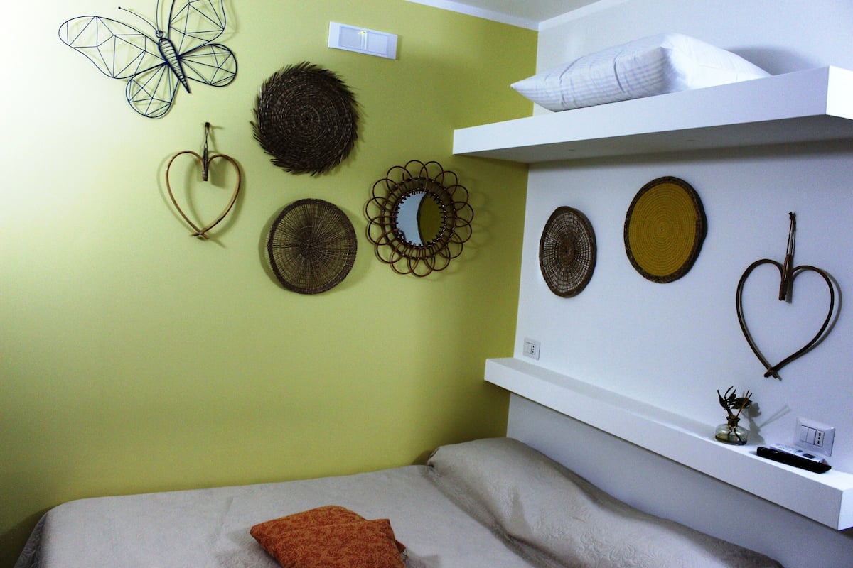 Ein Zimmer mit einem Bett, einem weißen Regal mit Dekorationen sowie Wandbildern, darunter ein Schmetterling, ein Herz und kreisförmige Muster.