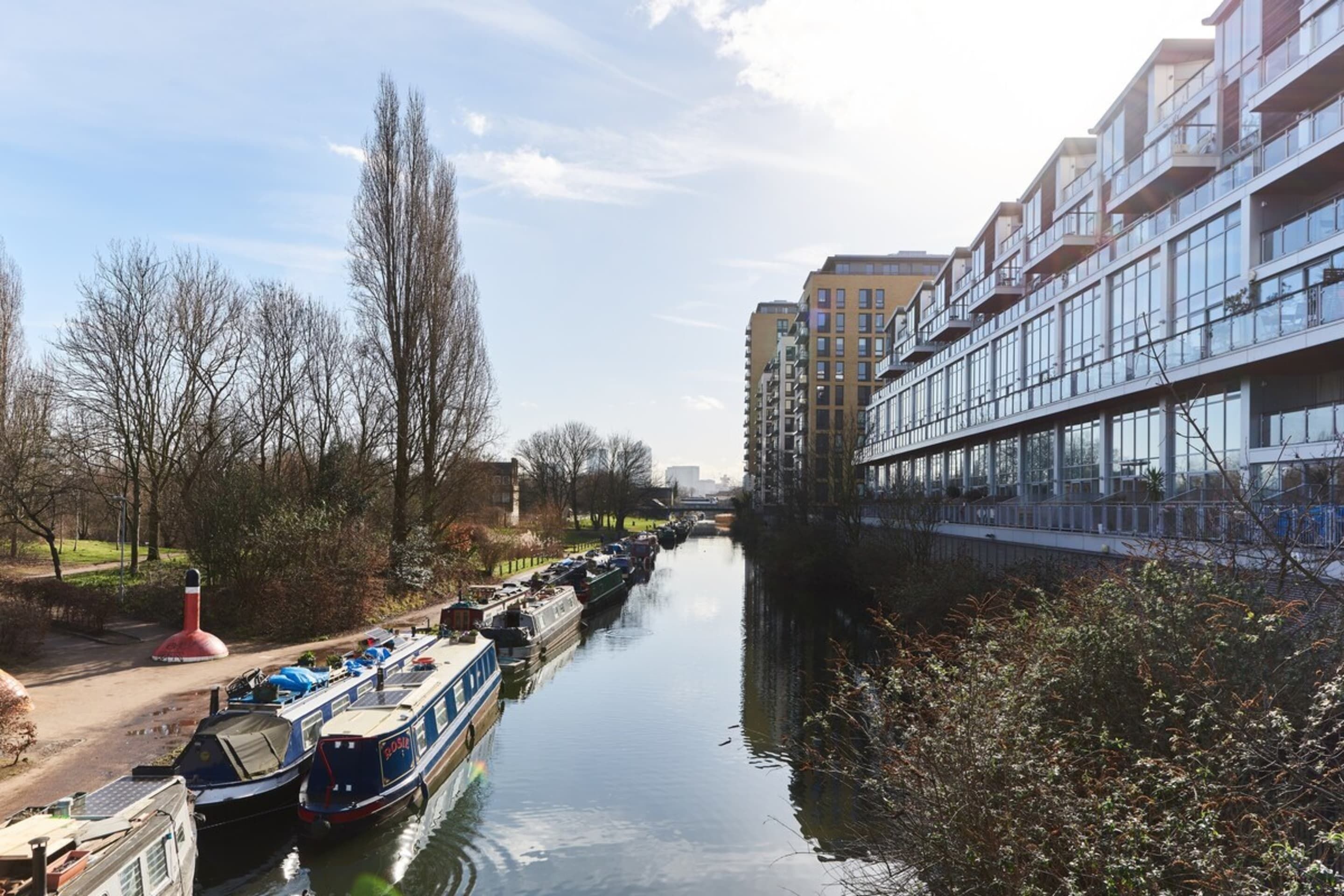 A narrow waterway with boats docked on the left, flanked by modern buildings and leafless trees.