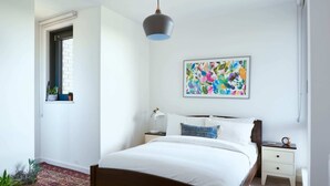 Una camera da letto con un letto, comodini, una lampada da comodino, una finestra con vista e un quadro astratto colorato appeso al muro.