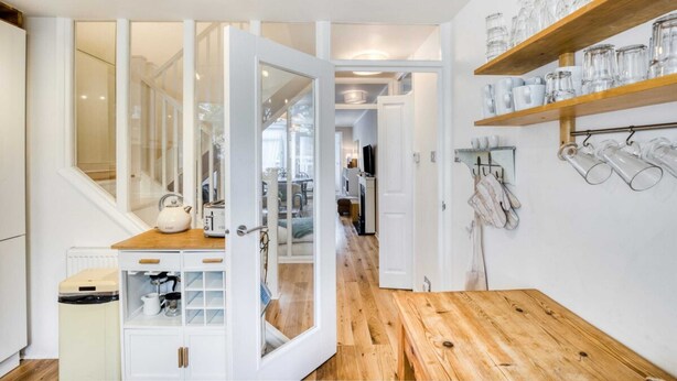 Une cuisine avec un plancher en bois, des meubles blancs et une porte en verre donnant sur une autre pièce.