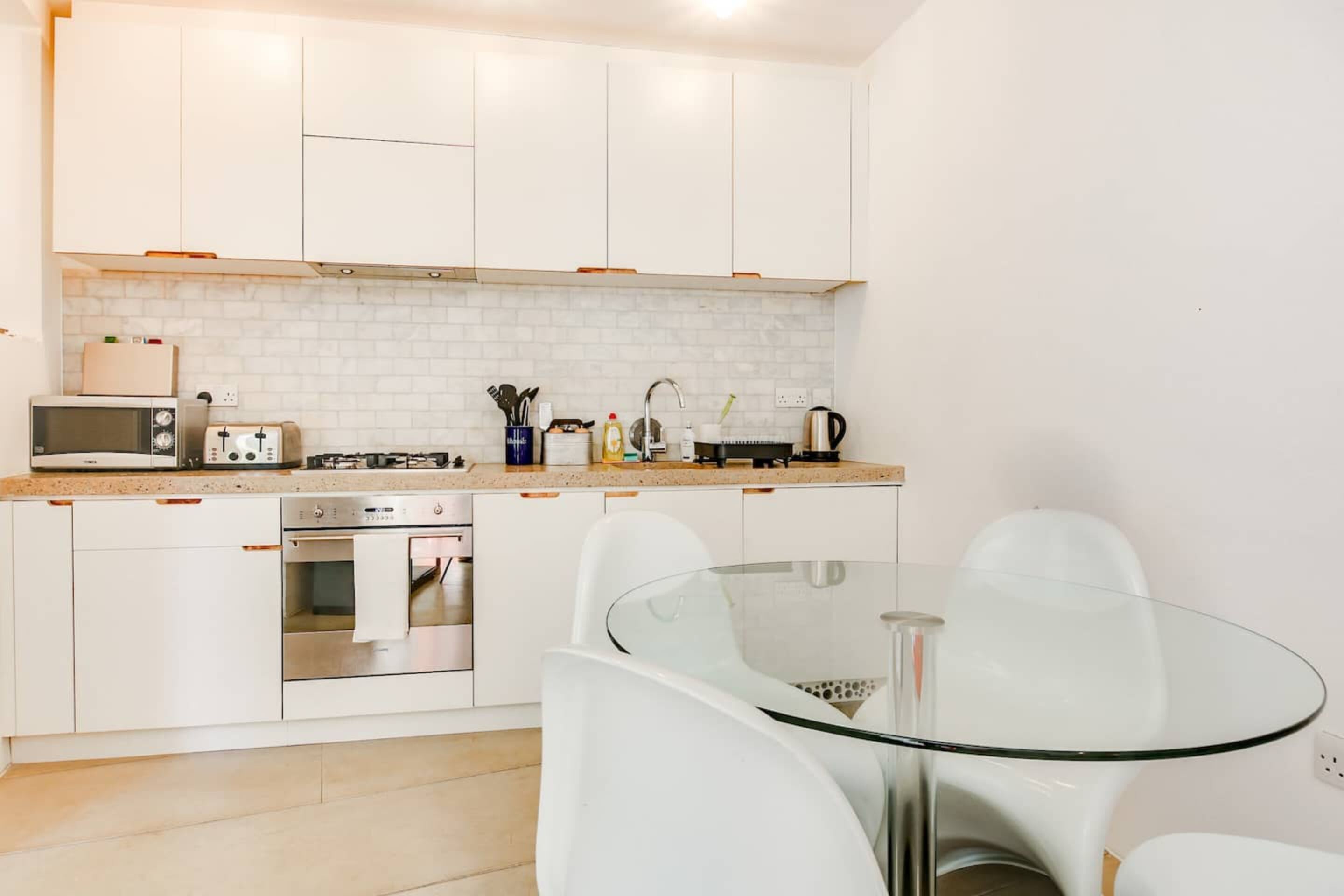 Una cocina moderna con armarios blancos, microondas, tostadora, estufa a gas y una mesa de comedor de vidrio con sillas.