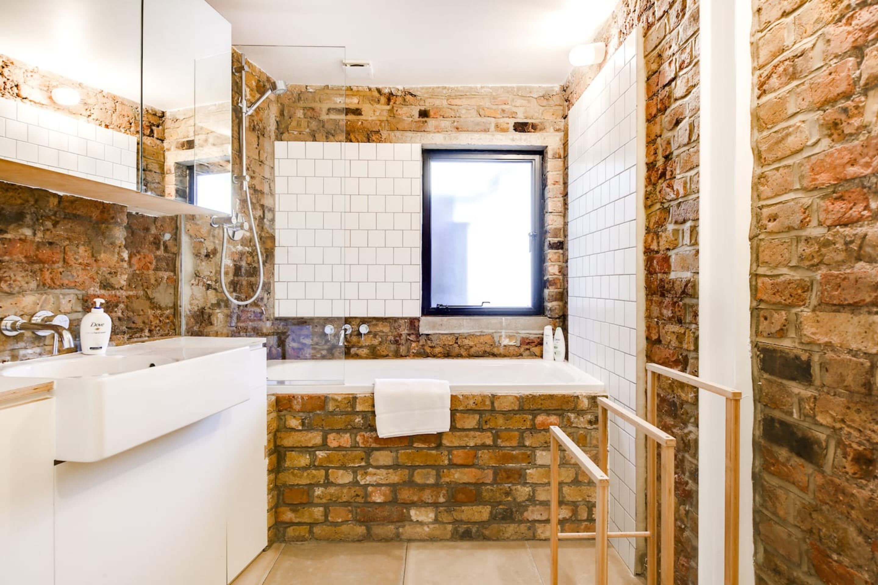 A bathroom with a brick wall, a window, a white sink, and a shower.