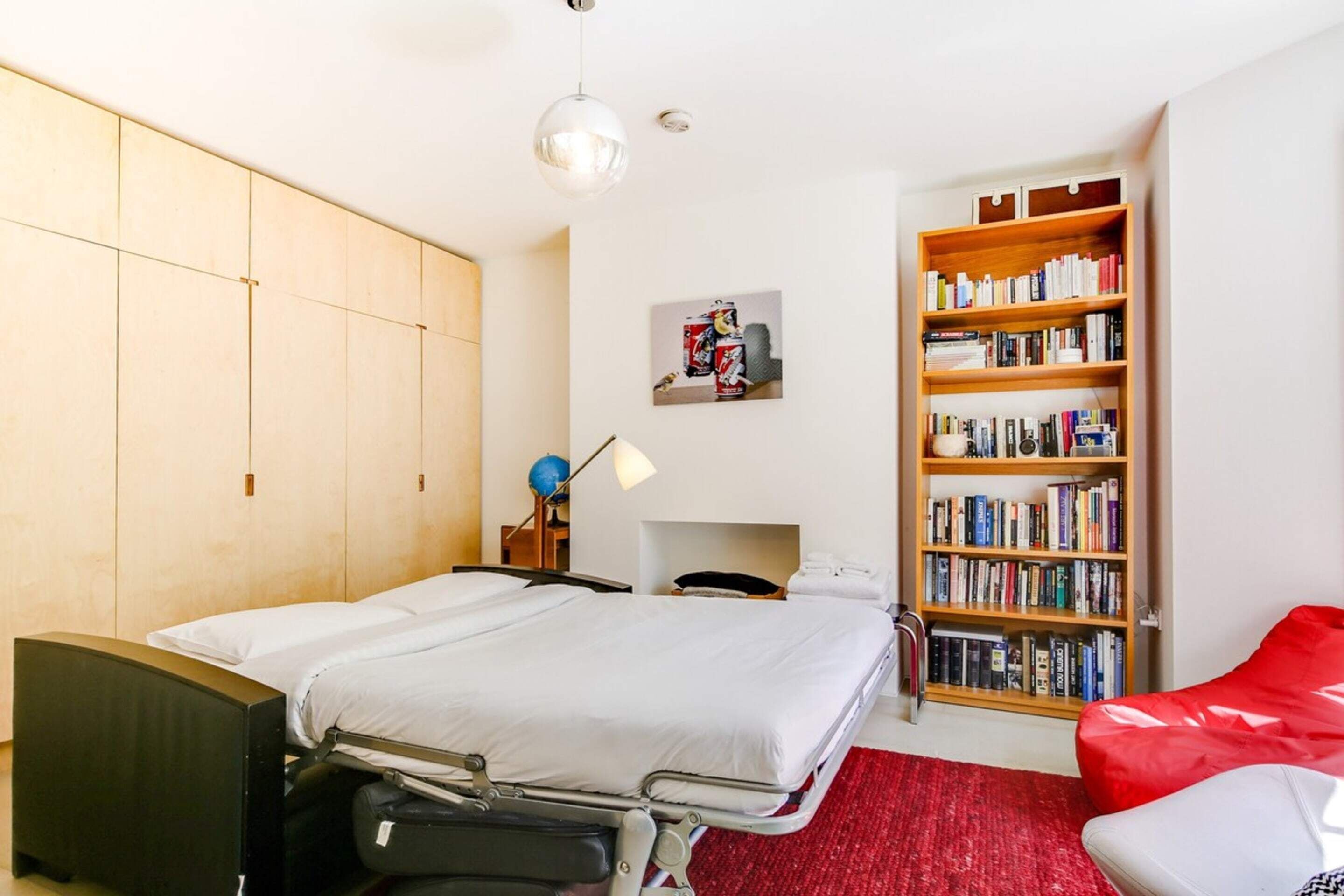 A bedroom with a bed, bedside table, lamp, bookshelf, and a red chair.