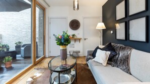 Un salon moderne avec une table basse en verre, un canapé et une grande fenêtre donnant sur un patio aménagé avec du mobilier d’extérieur.