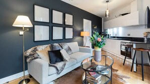 Un salon moderne avec un canapé, une table basse en verre et un lampadaire. On y trouve également un coin cuisine avec des meubles blancs et une table à manger avec des tabourets.