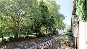 A balcony with a view of a tree-lined street, a parked car, and a building with a green awning.