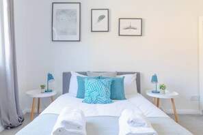 Een net opgemaakt bed met witte lakens en een blauw gestreept kussen, met aan weerszijden nachtkastjes met lampen en kleine plantjes.
