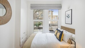 A bedroom with a bed, bedside table, lamp, and a window with a view of buildings.